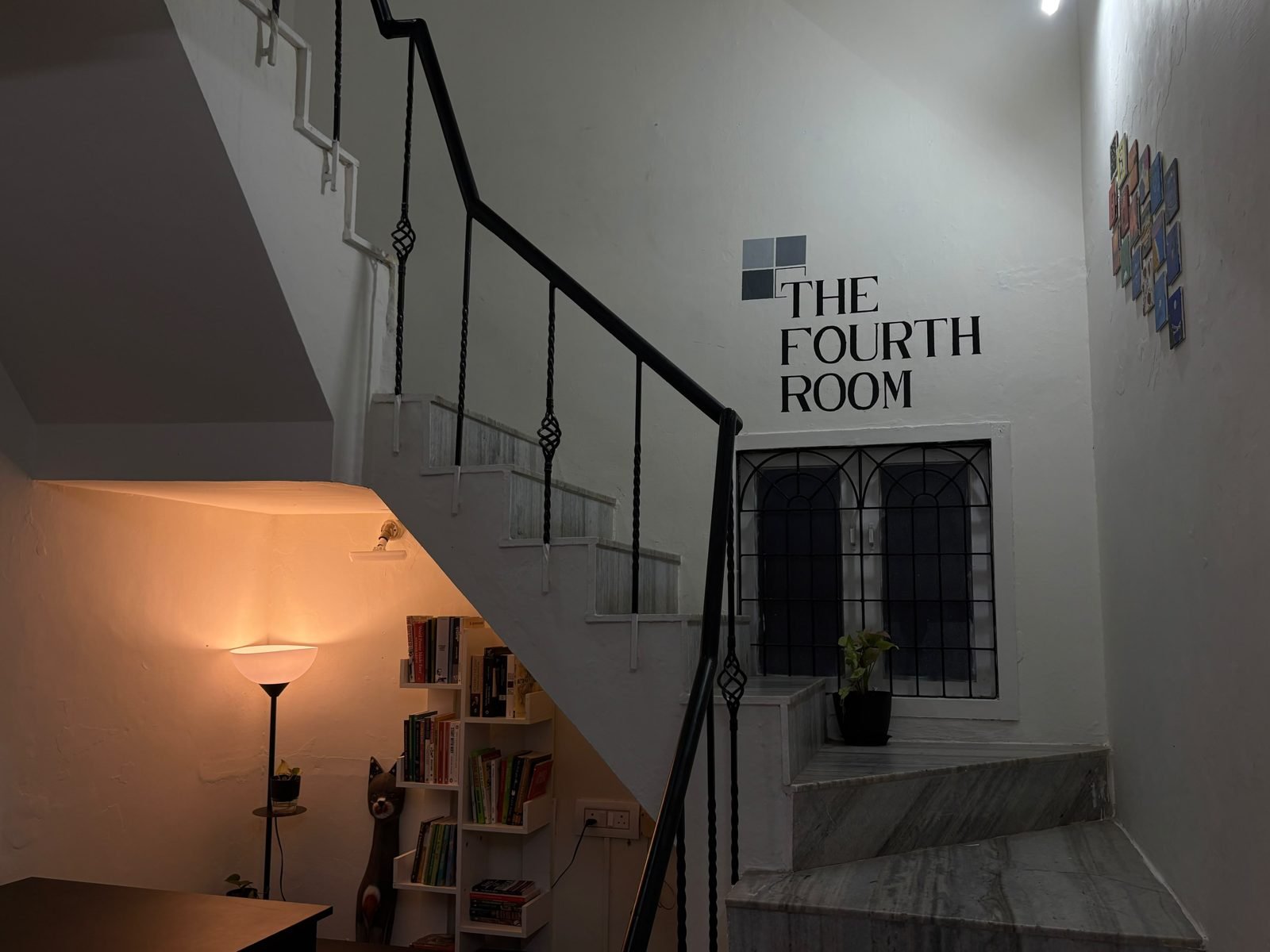 The ground-floor room in the evening: a floor lamp glows orange over a bookshelf in the nook under the stairs, beneath the painted sign 'The Fourth Room'.