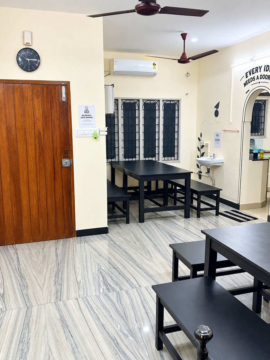 Rows of black tables and benches under ceiling fans and an air conditioner, with an arched doorway painted 'Every idea needs a door', a wall clock and a wash basin.